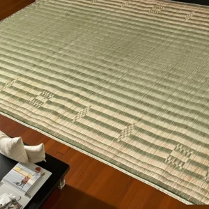 A handwoven 60x76 inch natural Korai grass floor mat with subtle traditional patterns, placed on a wooden floor near a black table and neutral decor.