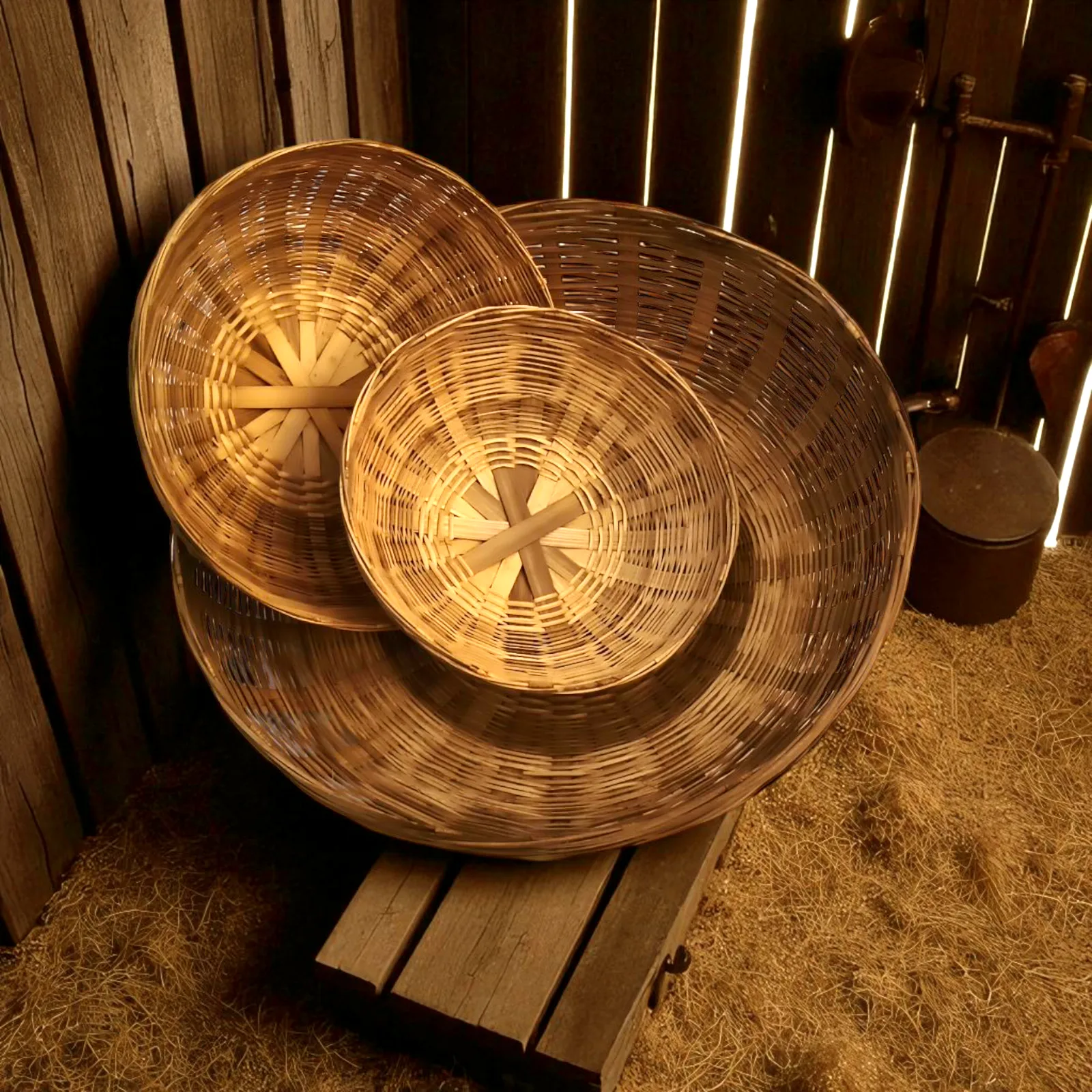 Three handwoven bamboo baskets placed inside a rustic wooden shed on a wooden stool with straw-covered flooring.