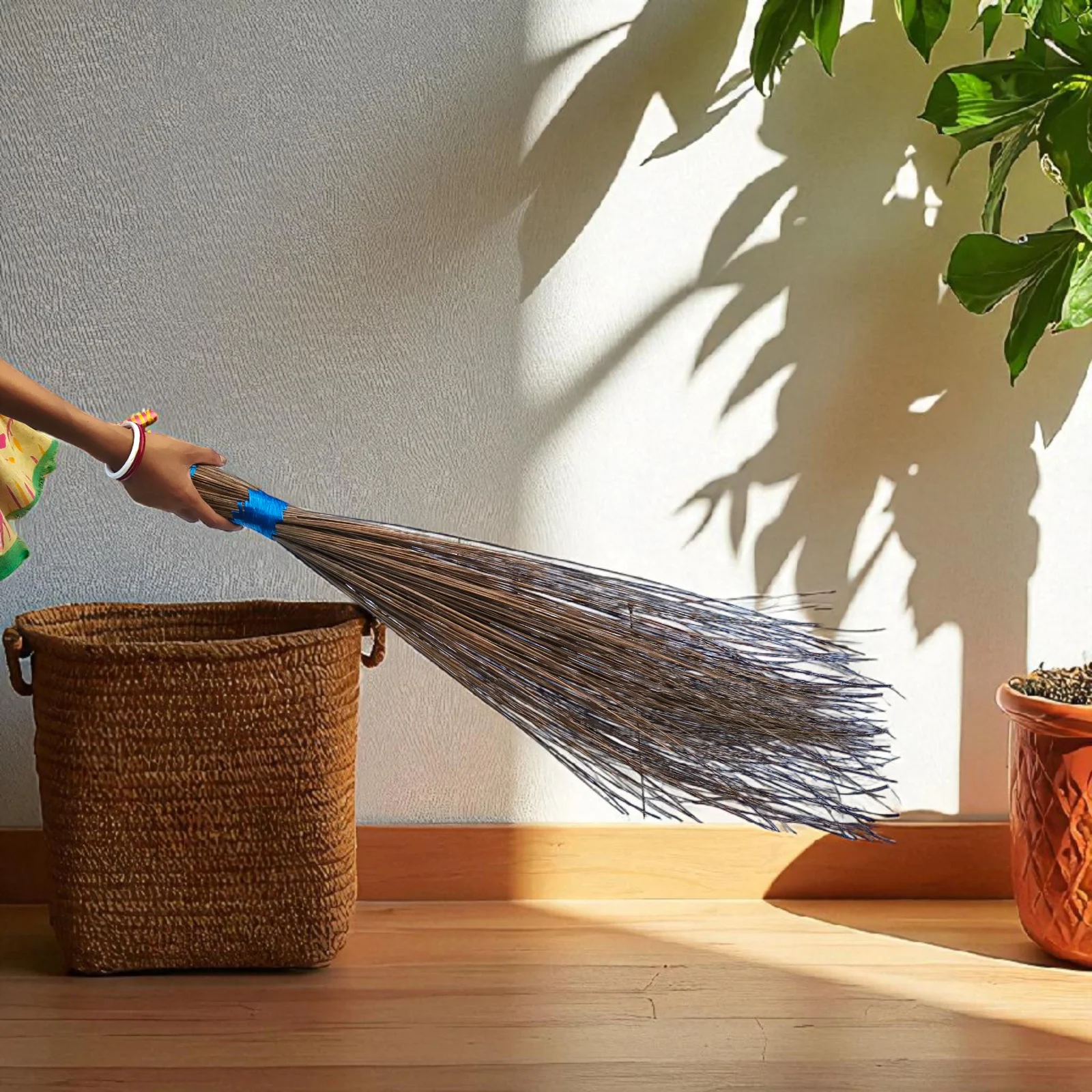 Handmade natural coconut broom with blue handle being used indoors beside a woven basket and potted plant.