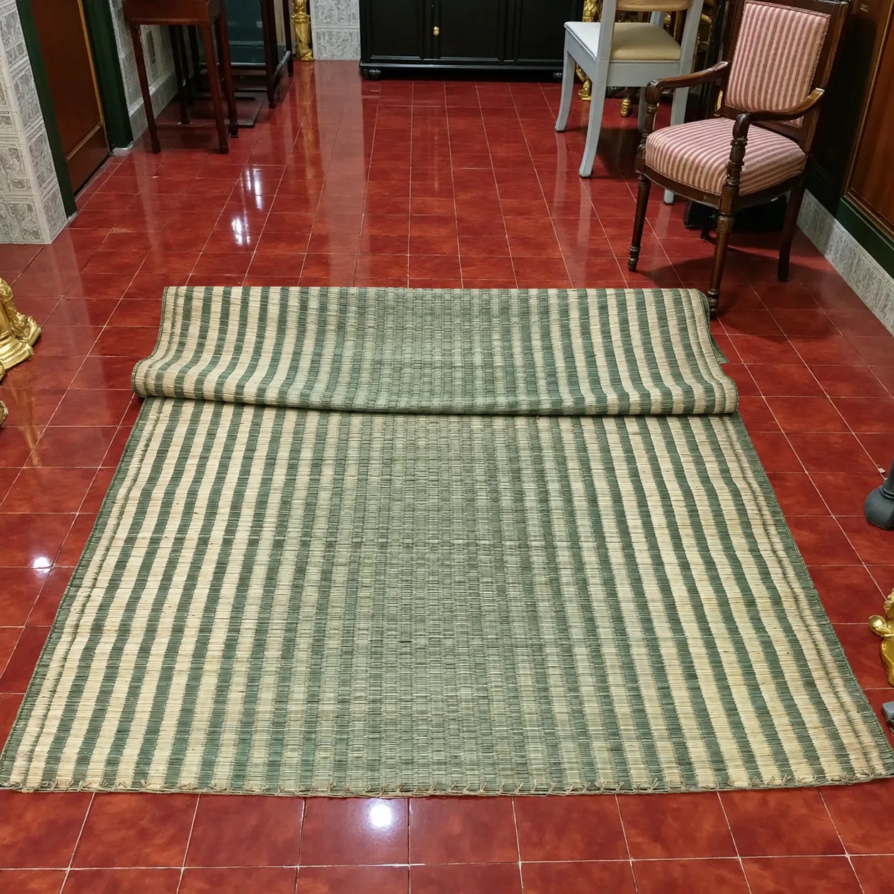 “Large handmade natural grass mat with green and cream striped woven design, partially rolled on a glossy red tile floor in a traditional home interior, showing its textured weave and finished edges.”